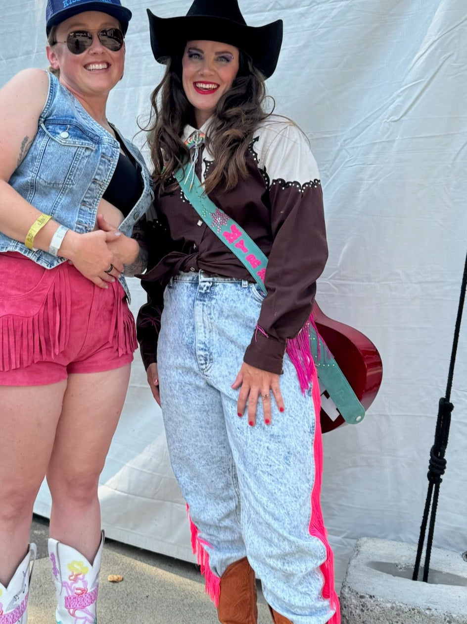 Two women posing together in front of a white tent, one in cowboy attire and the other in casual outfit with denim vest.